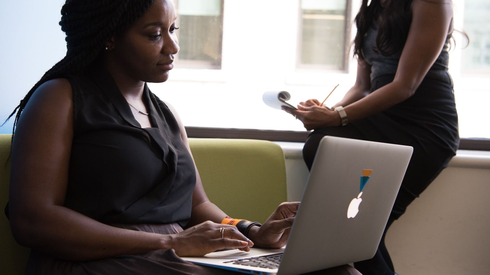 A woman using a laptop while a colleague stands beside her in an office.